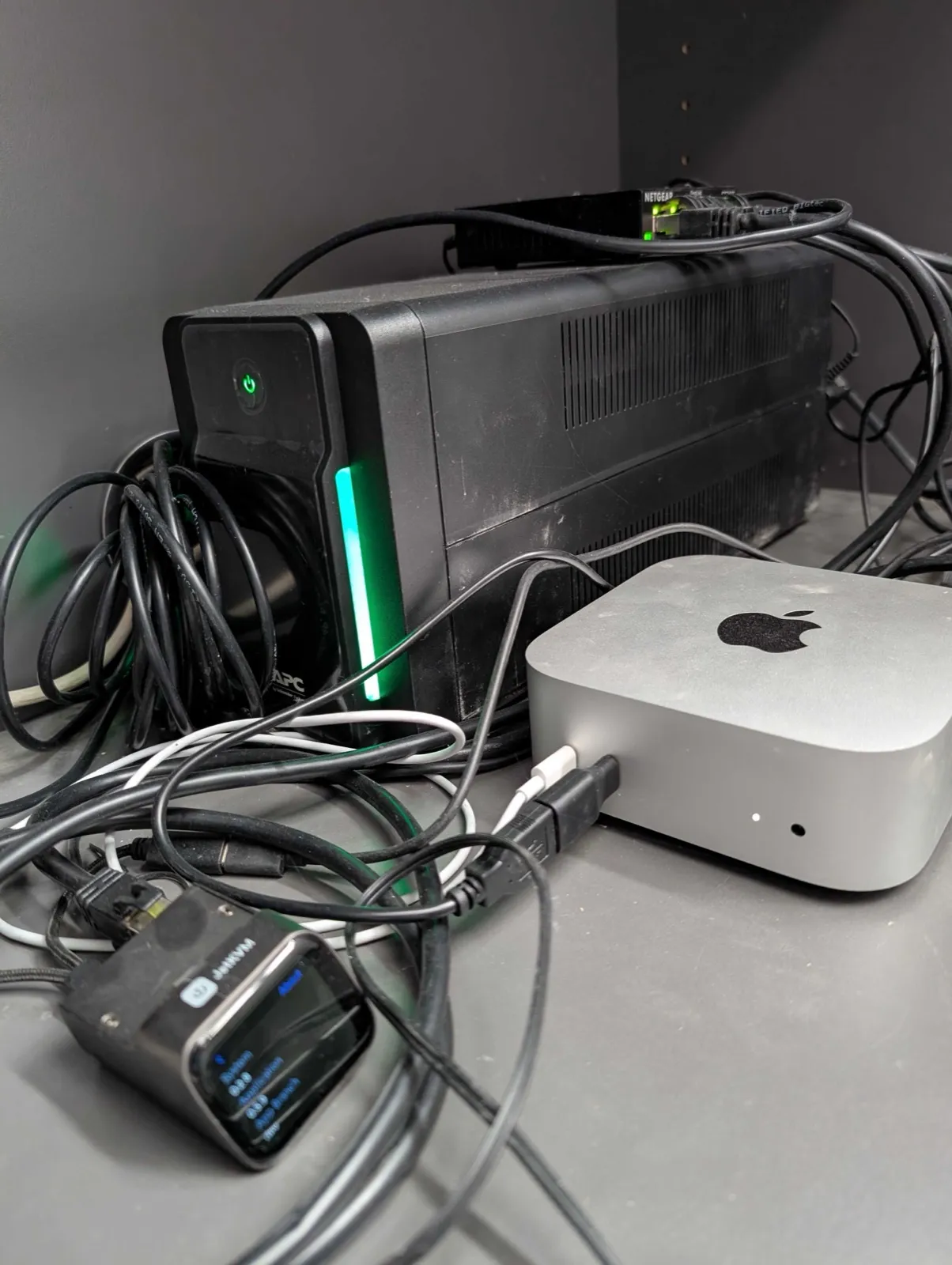 Metallregal mit der Hardware der Bodenstation: eine schwarze APC-USV mit grün leuchtender Statusleiste, davor ein silberner Mac mini, links unten ein JetKVM mit kleinem Farbdisplay, oben hinten ein NETGEAR-Switch mit grünen Port-LEDs, dazwischen zahlreiche schwarze und weiße Kabel.