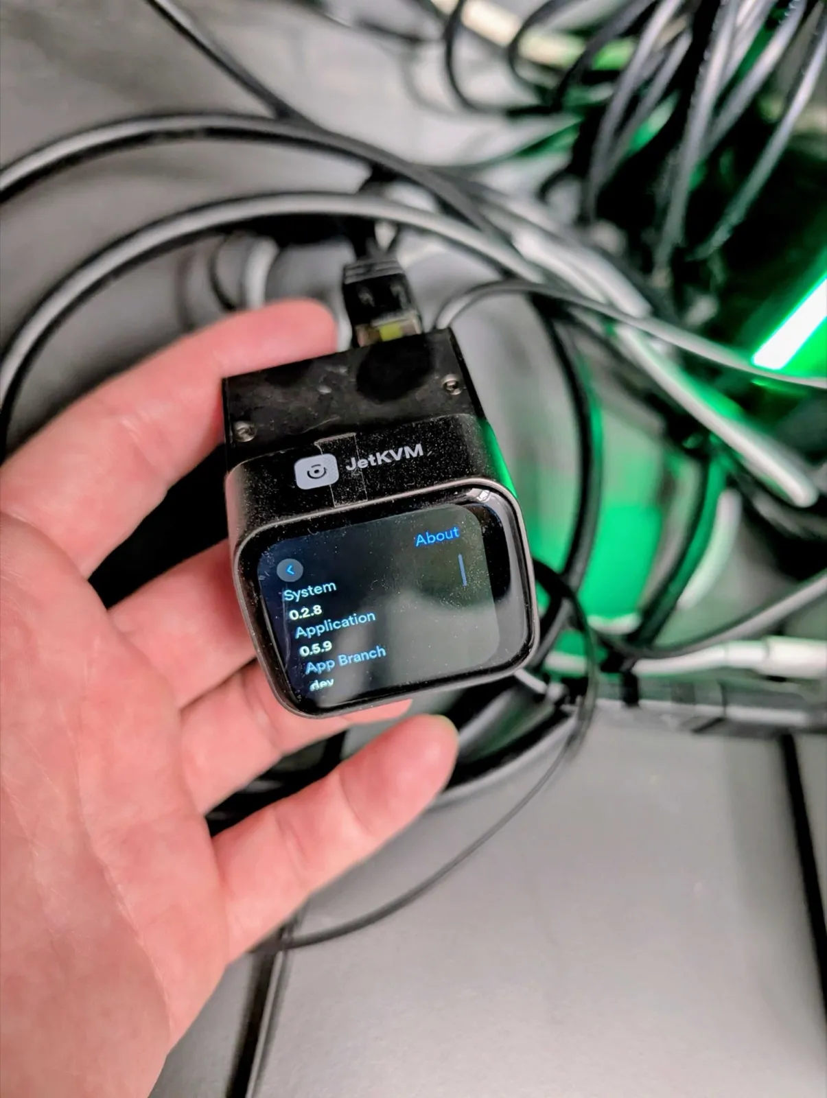 Foto: Eine Hand hält den JetKVM, ein kleines schwarzes KVM-over-IP-Gerät mit Farbdisplay, auf dem die Versions-Info steht (System 0.2.8, Application 0.5.9). Oben steckt ein Netzwerkkabel, im Hintergrund ein Kabelbündel in grünem Licht.